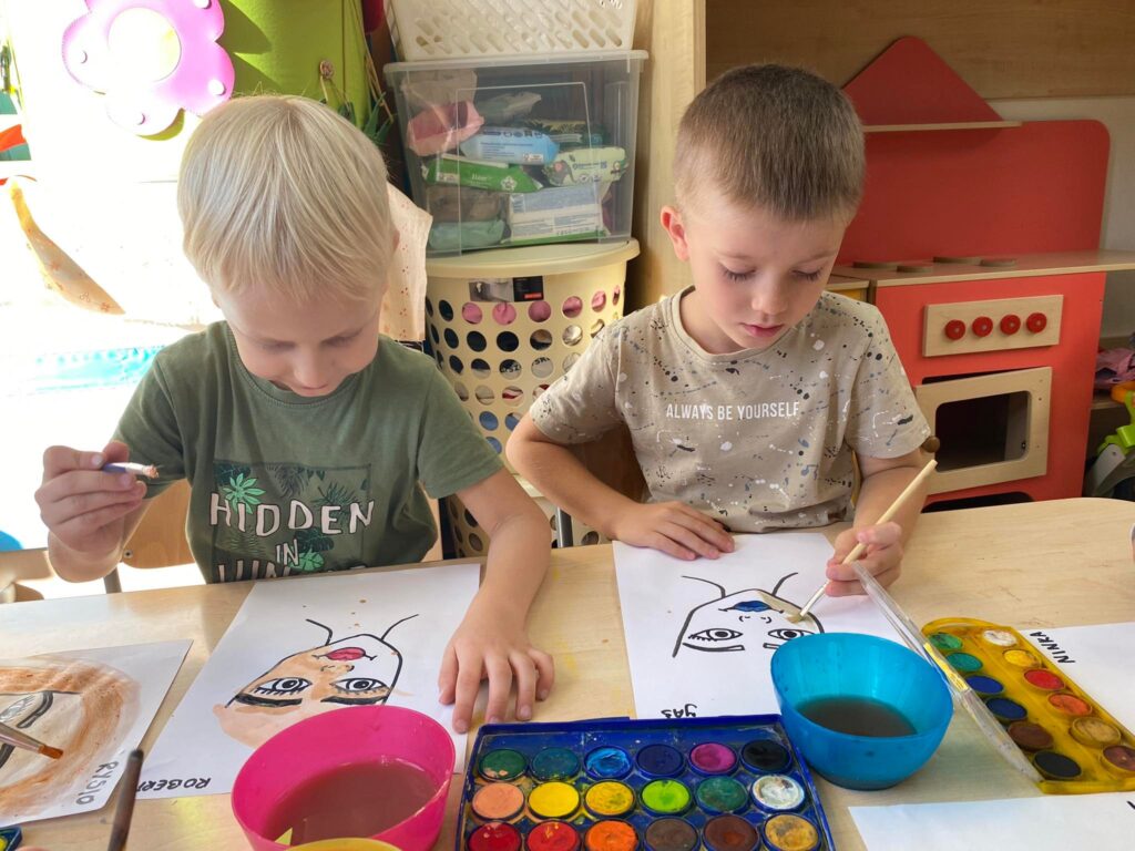 Dwóch chłopców siedząc przy stoliku malują farbami akwarelowymi.