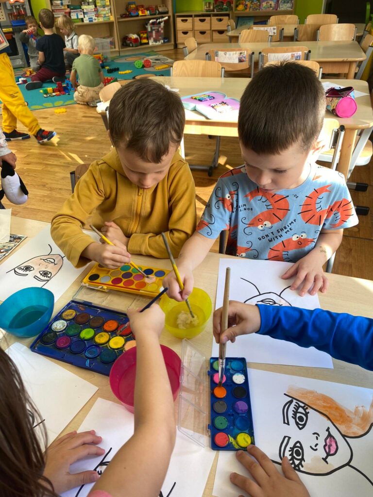Dwóch chłopców siedząc przy stoliku malują farbami akwarelowymi.