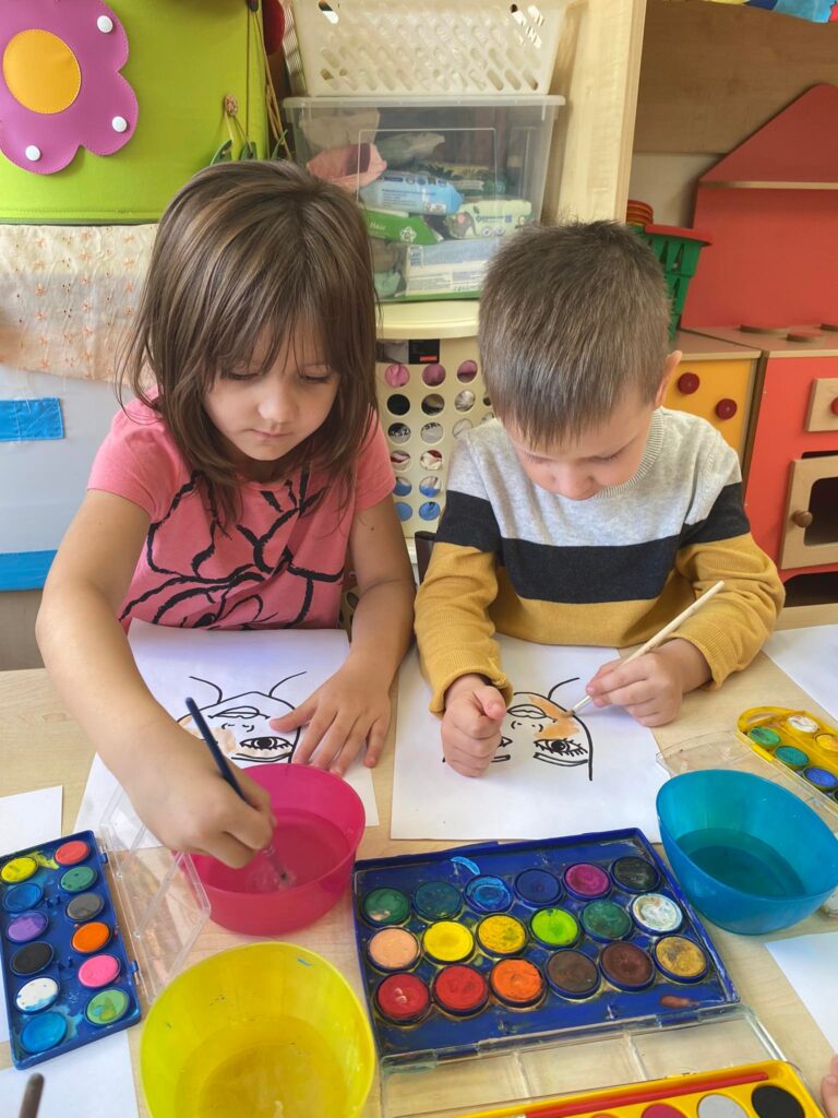 Dziewczynka i chłopiec siedząc przy stoliku malują farbami akwarelowymi.