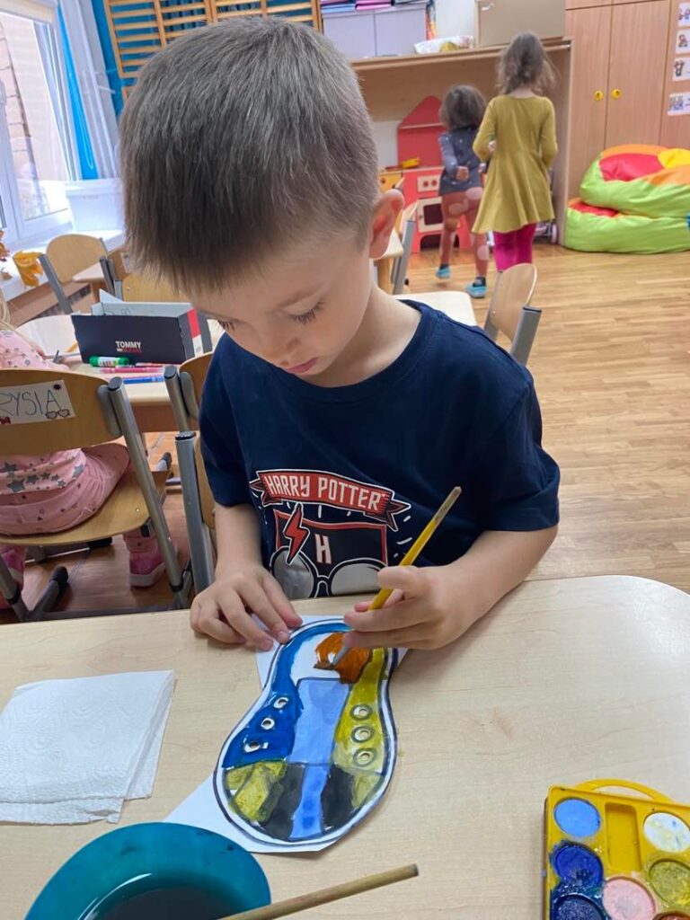 Chłopiec siedząc przy stoliku maluje farbami akwarelowymi papierowego trampka.