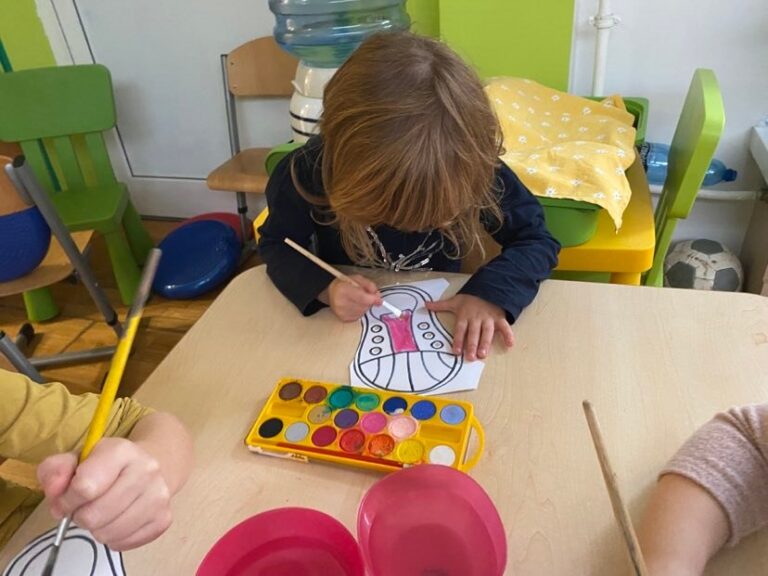 Dziewczynka siedząc przy stoliku maluje farbami akwarelowymi papierowego trampka.
