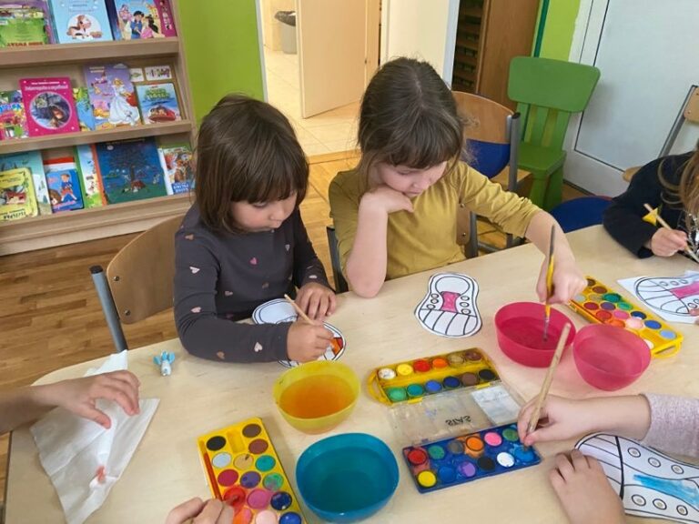 Dziewczynki siedząc przy stoliku malują farbami akwarelowymi papierowe trampki.