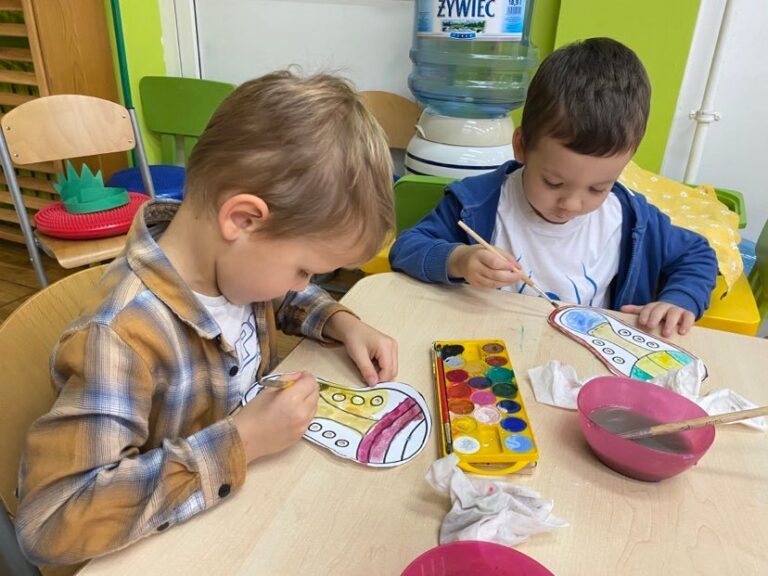 Chłopcy siedząc przy stoliku malują farbami akwarelowymi papierowe trampki.