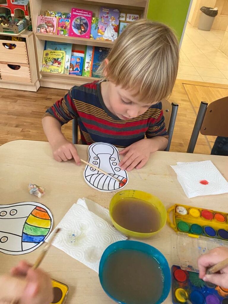 Chłopiec siedząc przy stoliku maluje farbami akwarelowymi papierowego trampka.