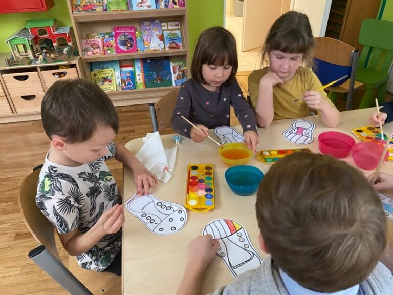Dzieci siedząc przy stoliku malują farbami akwarelowymi papierowe trampki.