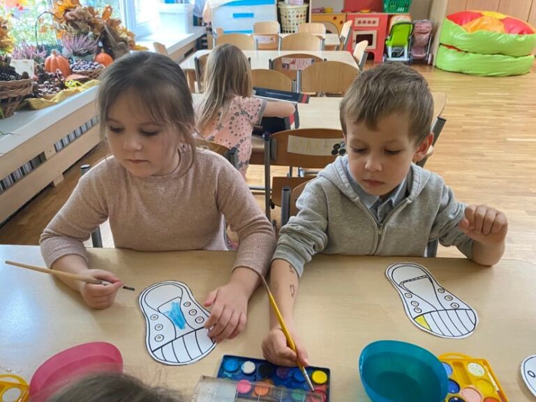 Dzieci siedząc przy stoliku malują farbami akwarelowymi papierowe trampki.