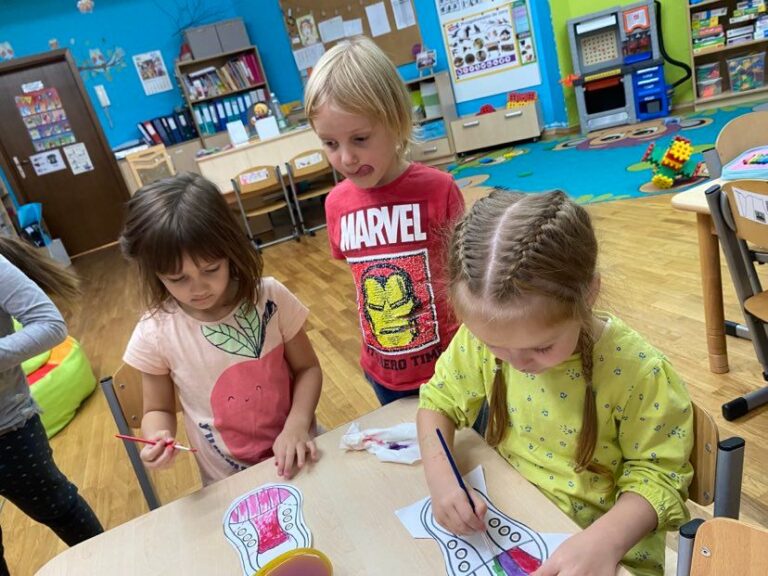 Dziewczynki siedząc przy stoliku malują farbami akwarelowymi papierowe trampki.