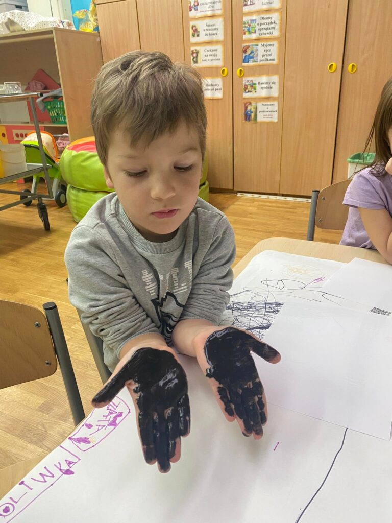 Przedszkolak siedząc przy stoliku pokazuje dłonie pomalowane czarną farbą.
