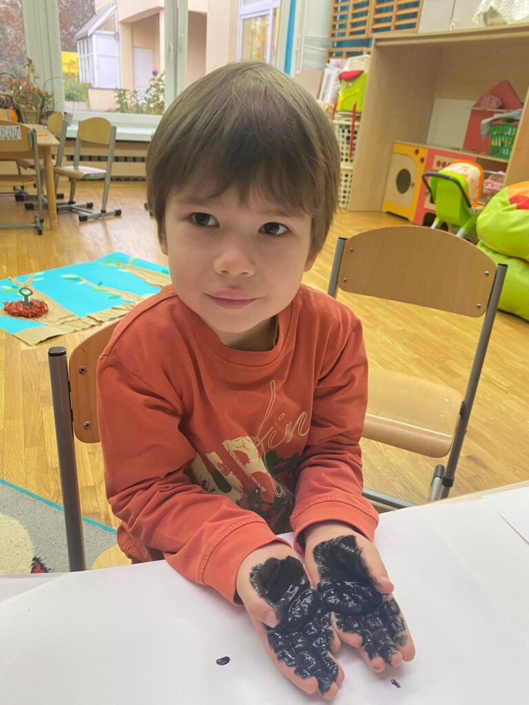 Przedszkolak siedząc przy stoliku pokazuje dłonie pomalowane czarną farbą.