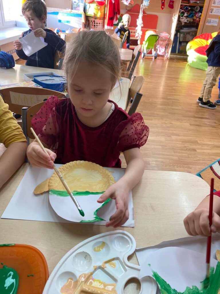 Dziewczynka siedząc przy stoliku maluje papierowe talerzyki za pomocą farb i pędzla.