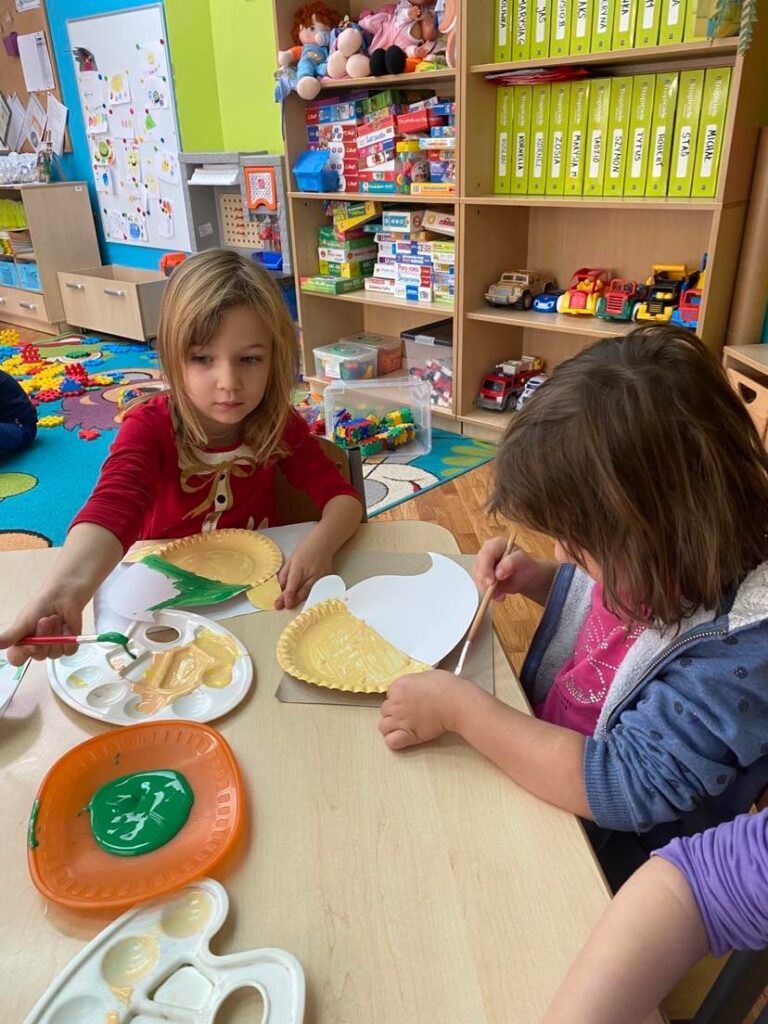 Dziewczynki siedząc przy stoliku malują papierowe talerzyki za pomocą farb i pędzla.
