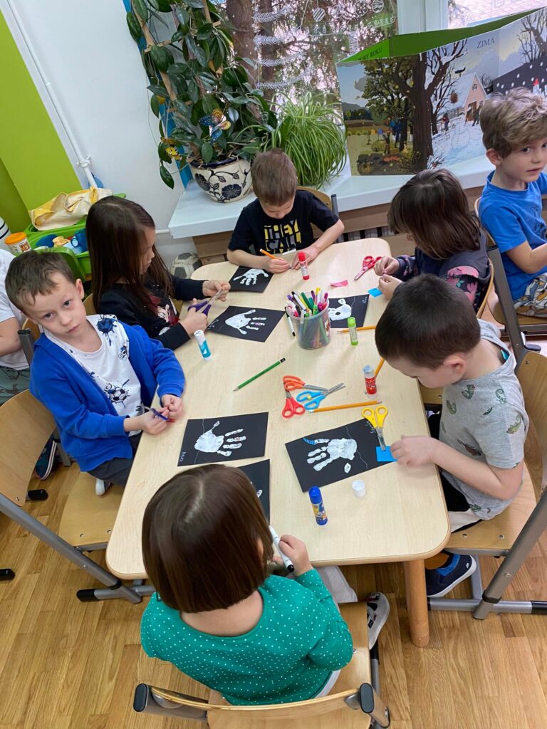 Dzieci siedząc przy stolikach wykonują prace plastyczną.