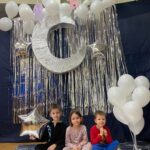 Przedszkolaki siedząc przy ściance z balonami, gwiazdami i księżycem pozują do pamiątkowego zdjęcia.