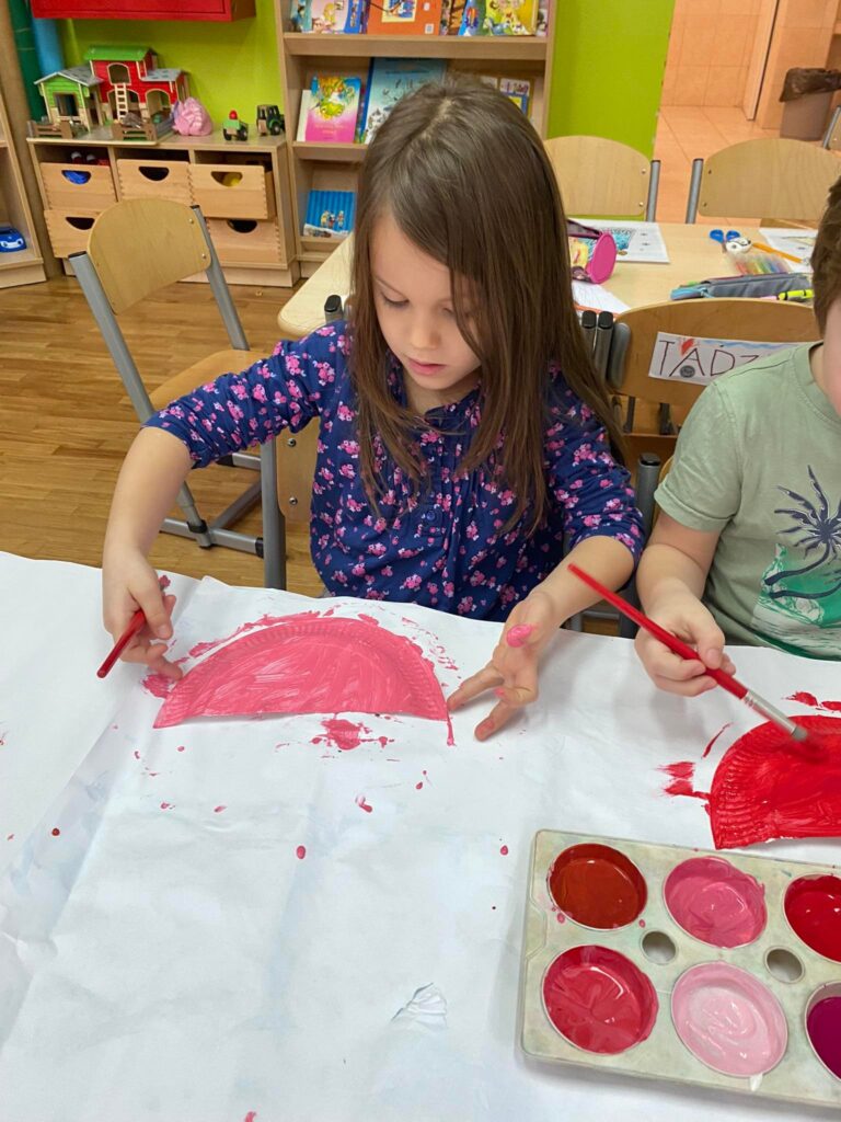 Dzieci siedząc przy stoliku malują farbami papierowe talerzyki.