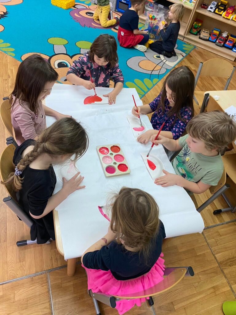 Dzieci siedząc przy stoliku malują farbami papierowe talerzyki.