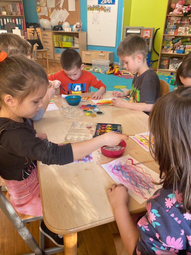 Dzieci siedząc przy stoliku malują farbami.