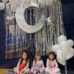 Przedszkolaki siedząc przy ściance z balonami, gwiazdami i księżycem pozują do pamiątkowego zdjęcia.