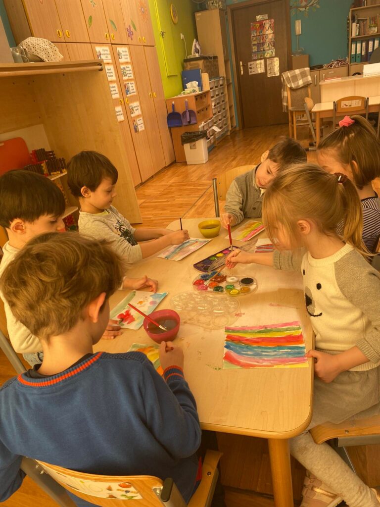 Dzieci siedząc przy stoliku malują farbami.