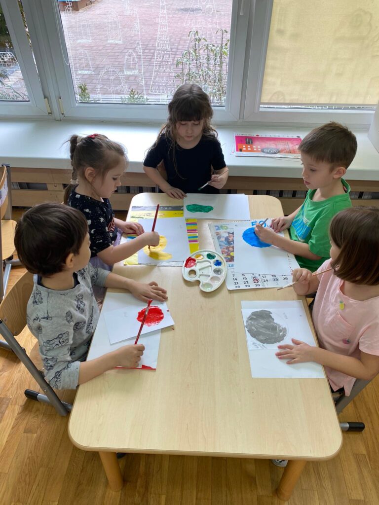 Dzieci siedząc przy stoliku malują papierowe planety.