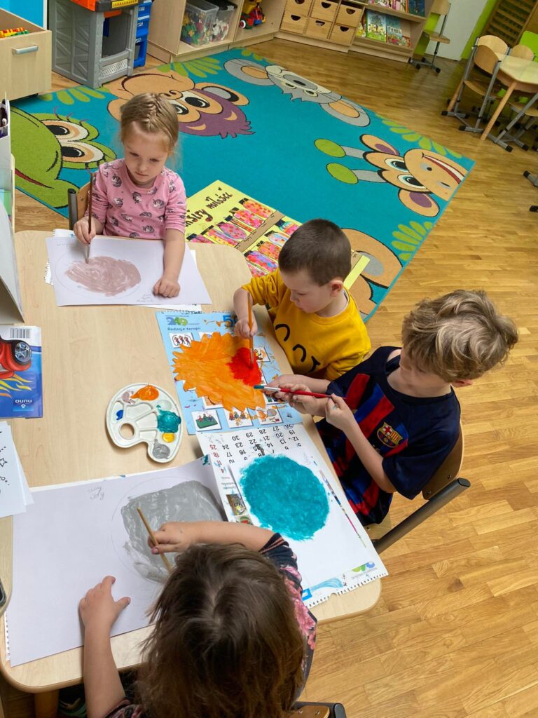 Dzieci siedząc przy stoliku malują papierowe planety.