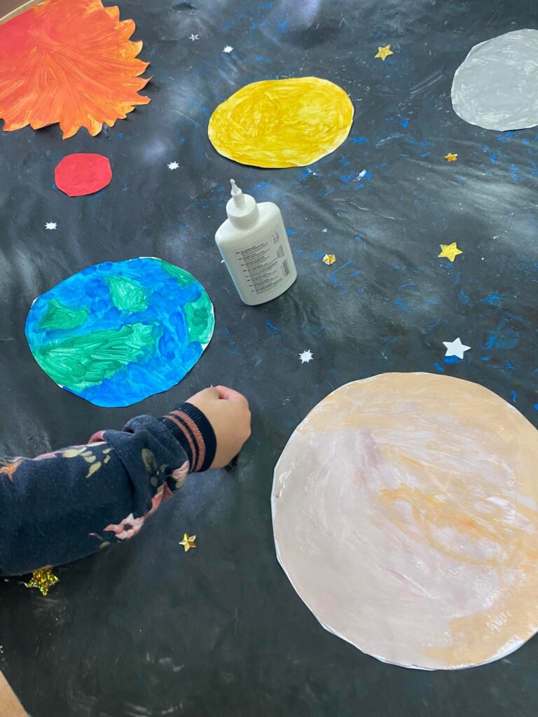 Dziewczynka przykleja gwiazdy do brystolu z namalowanym kosmosem.