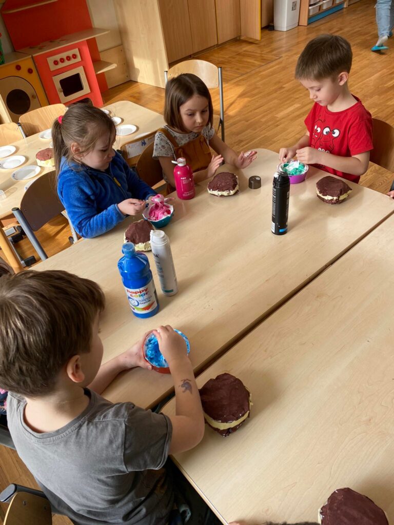 Dzieci siedząc przy stoliku przygotowują polewę do papierowych pączków.