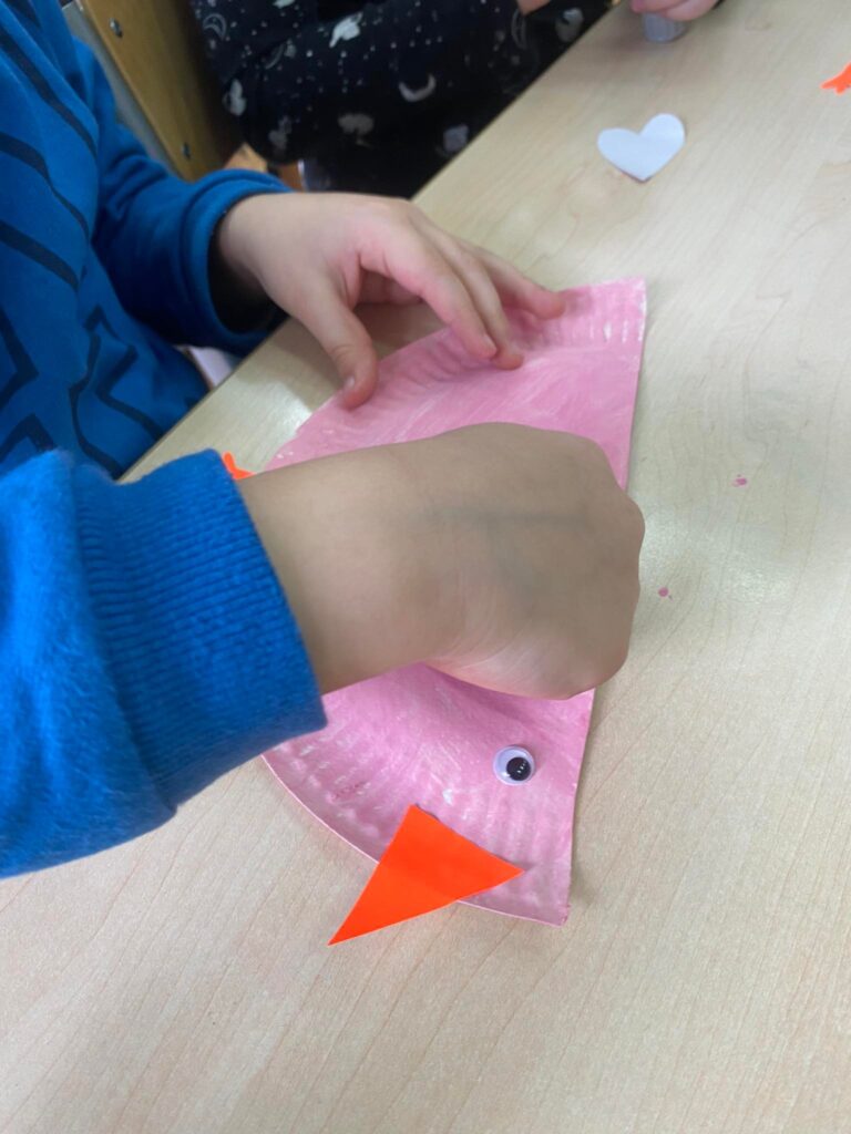 Dziecko przykleja papierowe skrzydełko do koguta zrobionego z talerzyka.