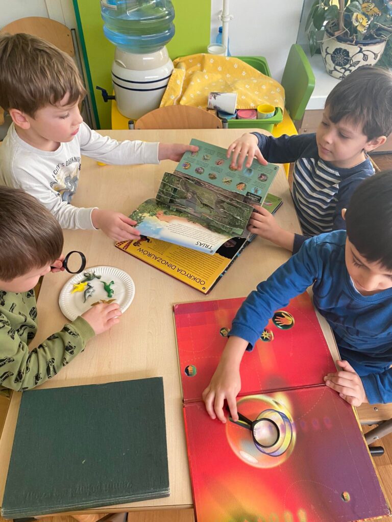 Dzieci oglądają książki o dinozaurach oraz oglądają przez lupę figurki dinozaurów.