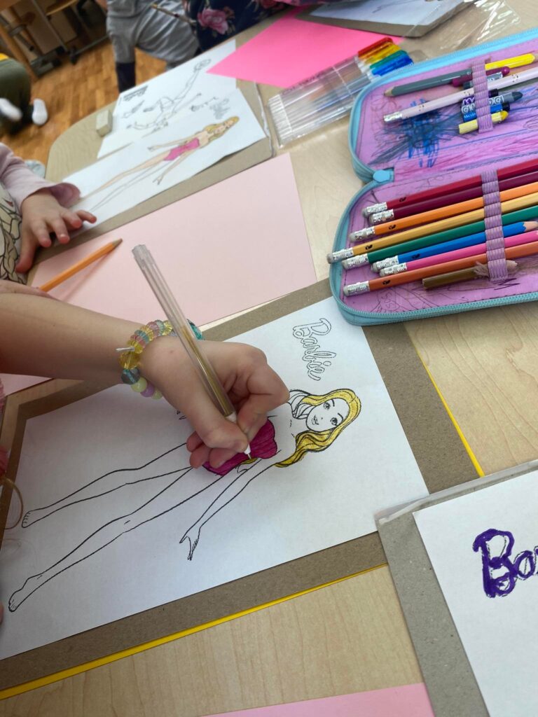 Dziewczynki wykonują prace plastyczne przedstawiającą Barbie w sukience.