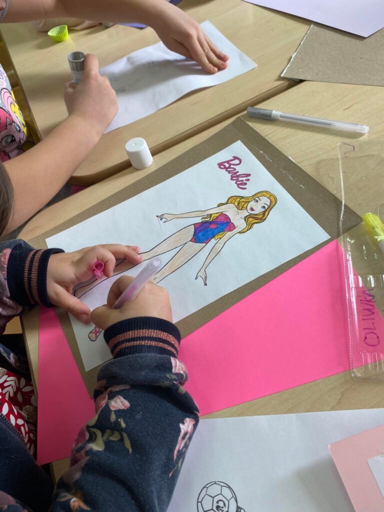 Dziewczynki wykonują prace plastyczne przedstawiającą Barbie w sukience.