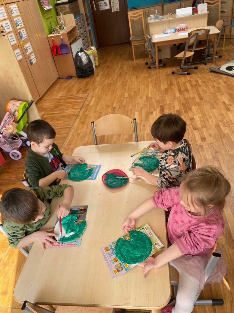 Dzieci siedząc przy stolikach malują papierowe talerzyki na zielono.