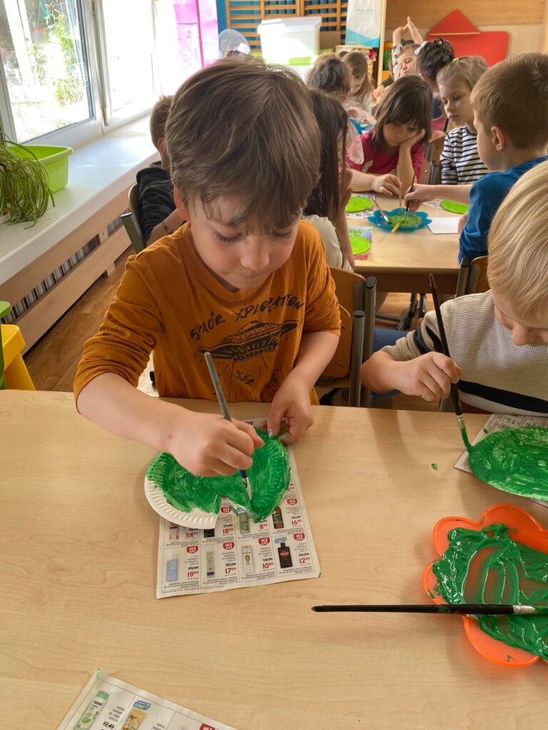 Chłopiec siedząc przy stoliku maluje papierowy talerzyk na zielono.