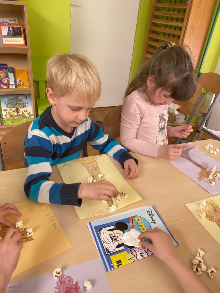 Przedszkolaki siedząc przy stoliku przyklejają popcorn do narysowanych przez siebie drzew.