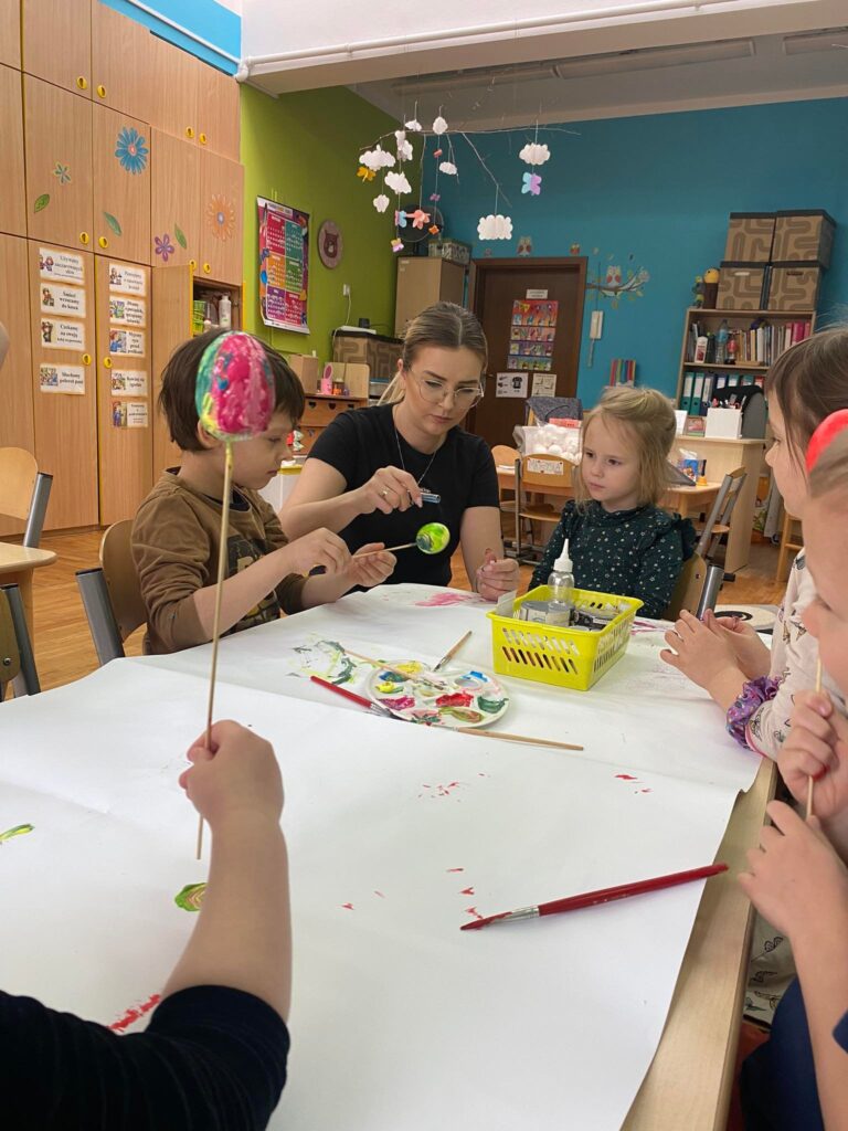 Dzieci razem z nauczycielem siedząc przy stoliku malują styropianowe jajka farbą.