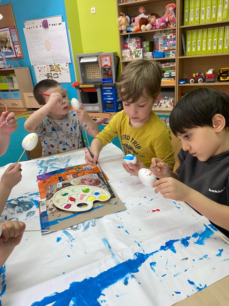 Dzieci siedząc przy stoliku malują styropianowe jajka farbą.