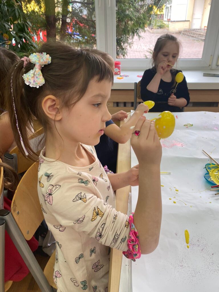 Dzieci siedząc przy stoliku malują styropianowe jajka farbą.