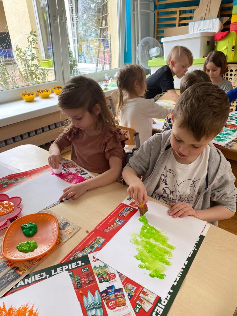 Dzieci siedząc przy stoliku malują kartki farbami metodą stemplowania.