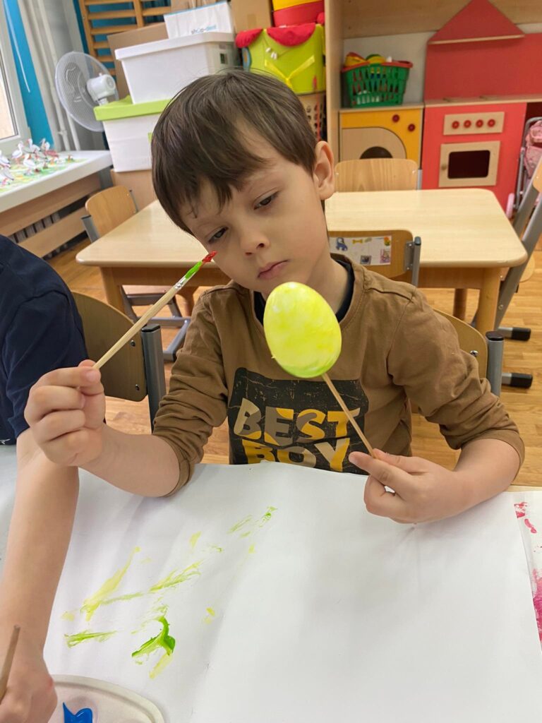 Dziecko siedząc przy stoliku maluje styropianowe jajko farbą.