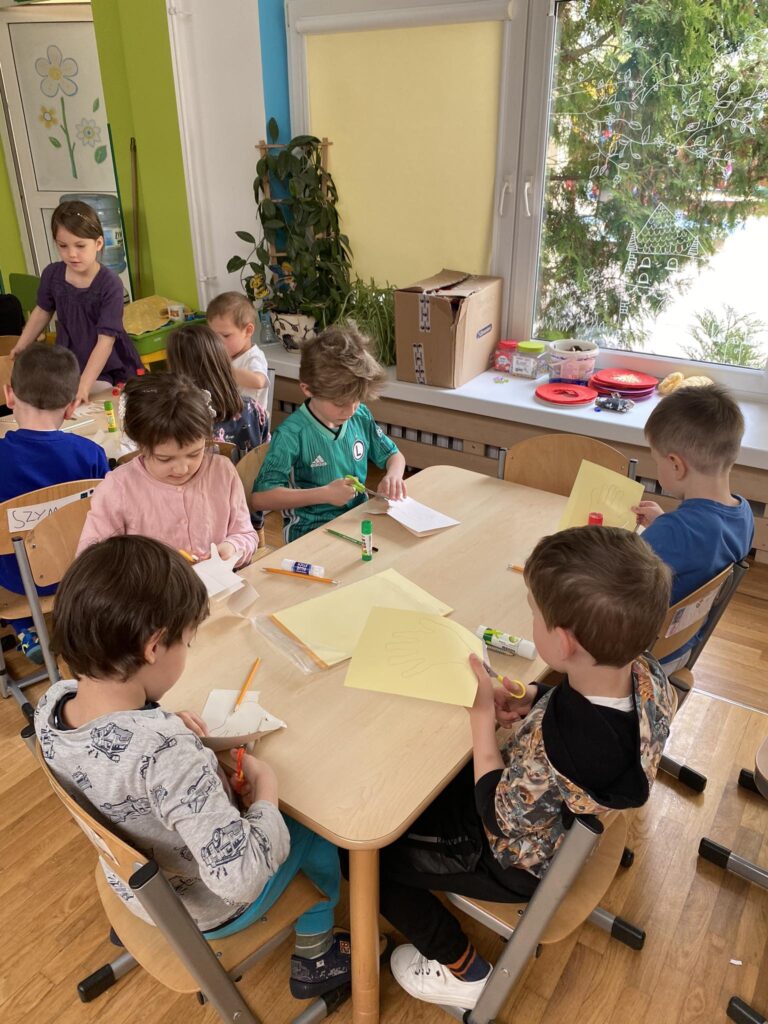 Dzieci siedząc przy stoliku wykonują prace plastyczną.