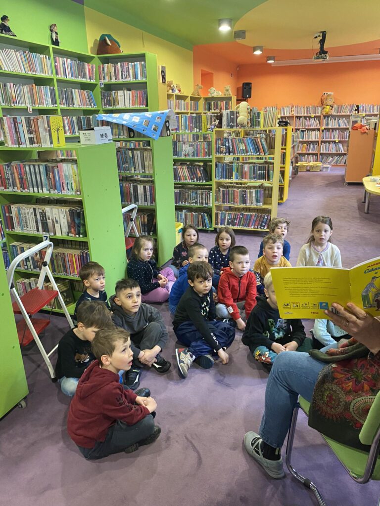 Dzieci siedząc na dywanie biorą udział w warsztatach bibliotecznych.