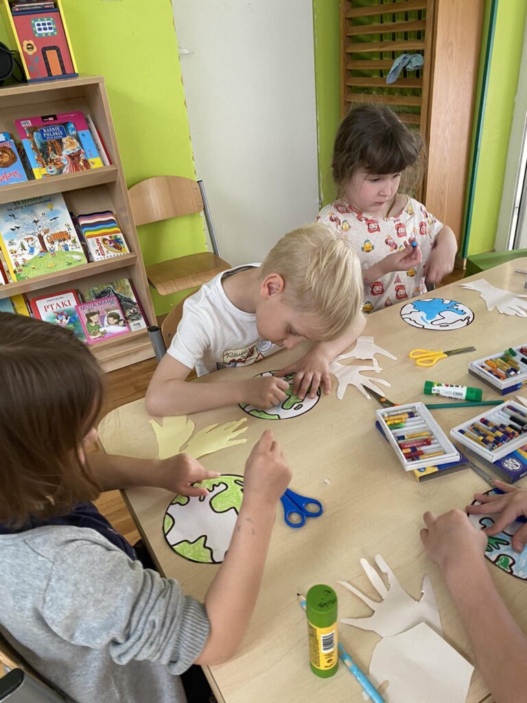 Dzieci siedząc przy stoliku wykonują prace plastyczną.