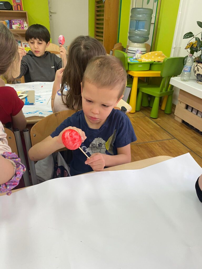 Dziecko siedząc przy stoliku maluje styropianowe jajko farbą.