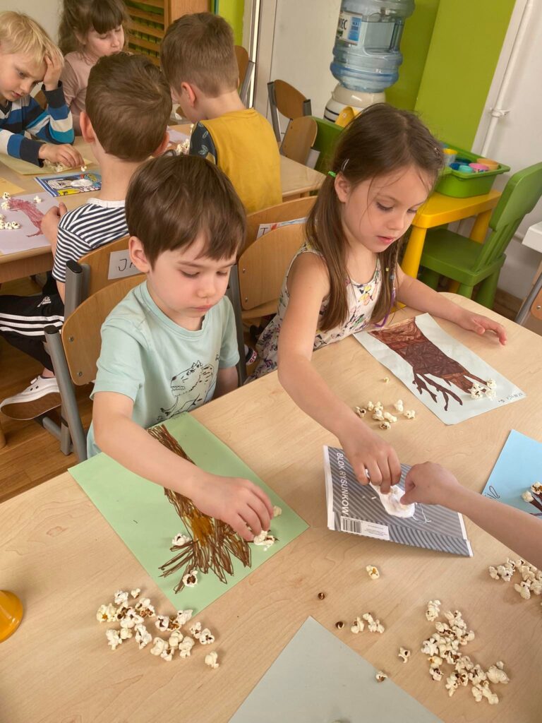 Przedszkolaki siedząc przy stoliku przyklejają popcorn do narysowanych przez siebie drzew.